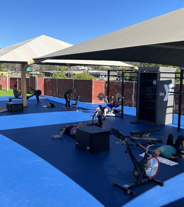 People exercise on mats and stationary bikes under shade structures in the updated gym workout space at Marin YMCA, with fencing and trees in the background on a sunny day.