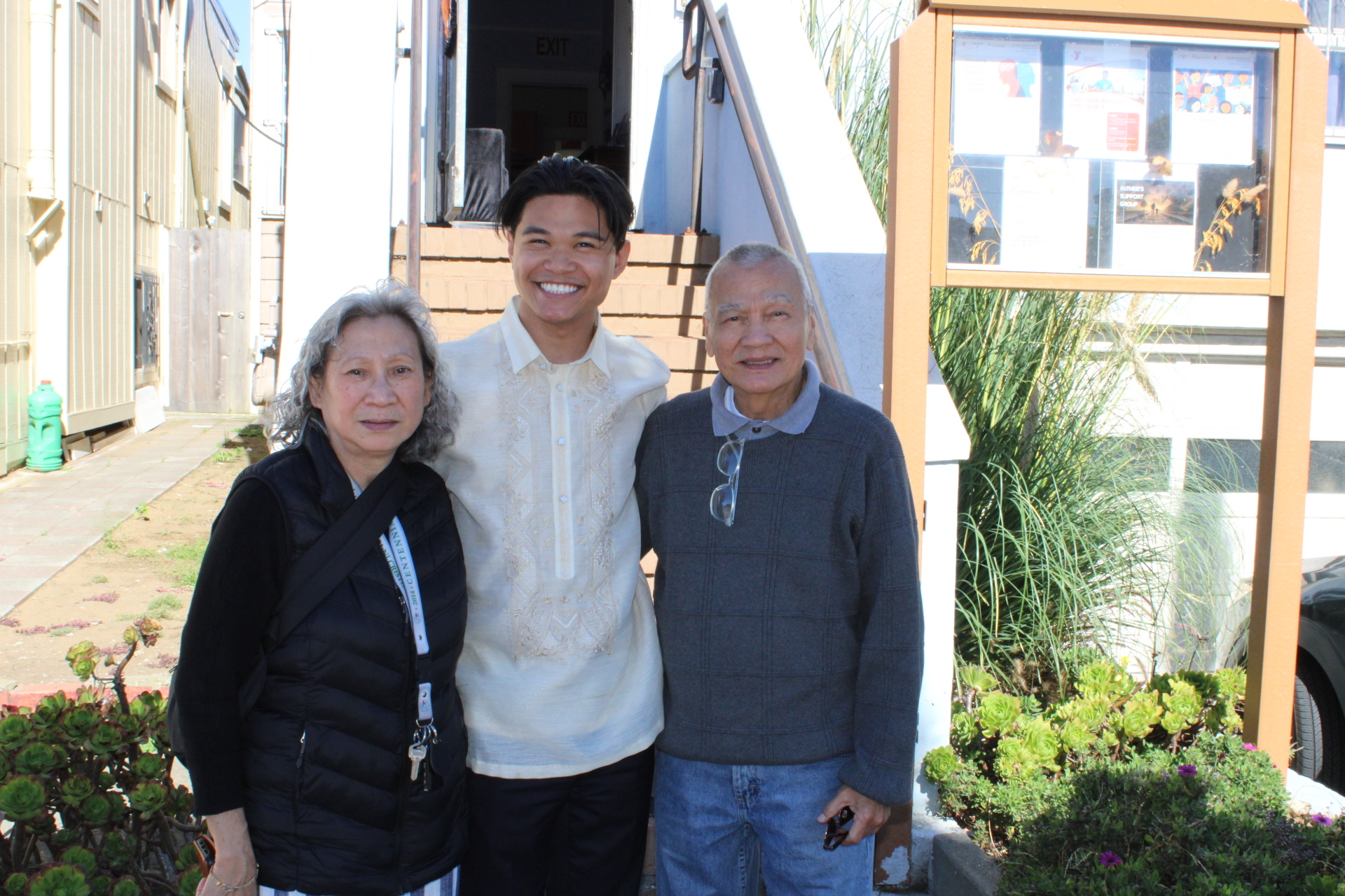 Three people are standing outdoors in front of stairs, smiling at the camera. The person in the middle wears a traditional barong tagalog, while the others wear casual clothes, enjoying a moment of wellness near the Marin YMCA.