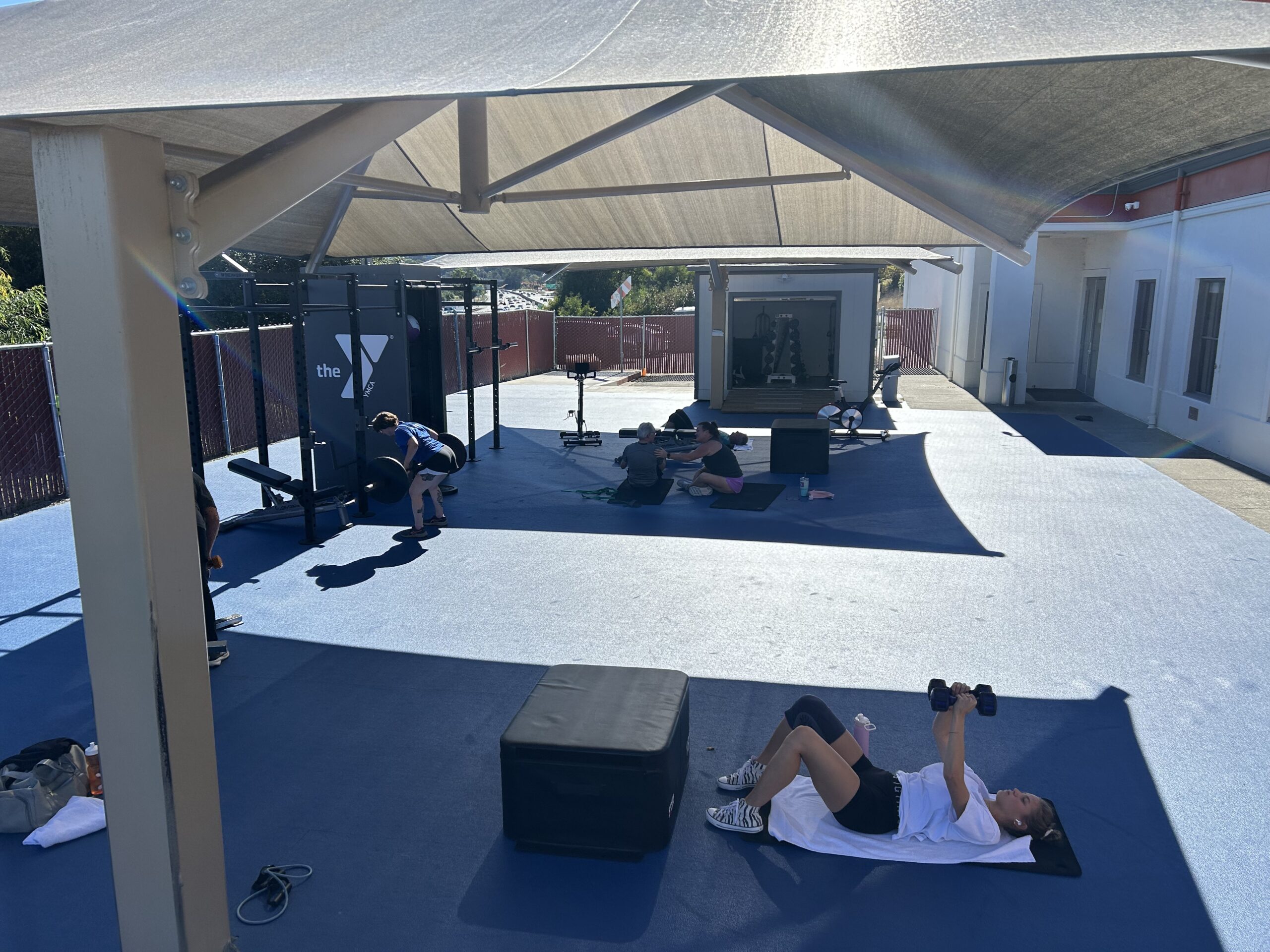 Several people exercise in a vibrant outdoor workout space at the Marin YMCA, using weights and resistance bands on blue flooring under a large canopy, promoting wellness near the building.