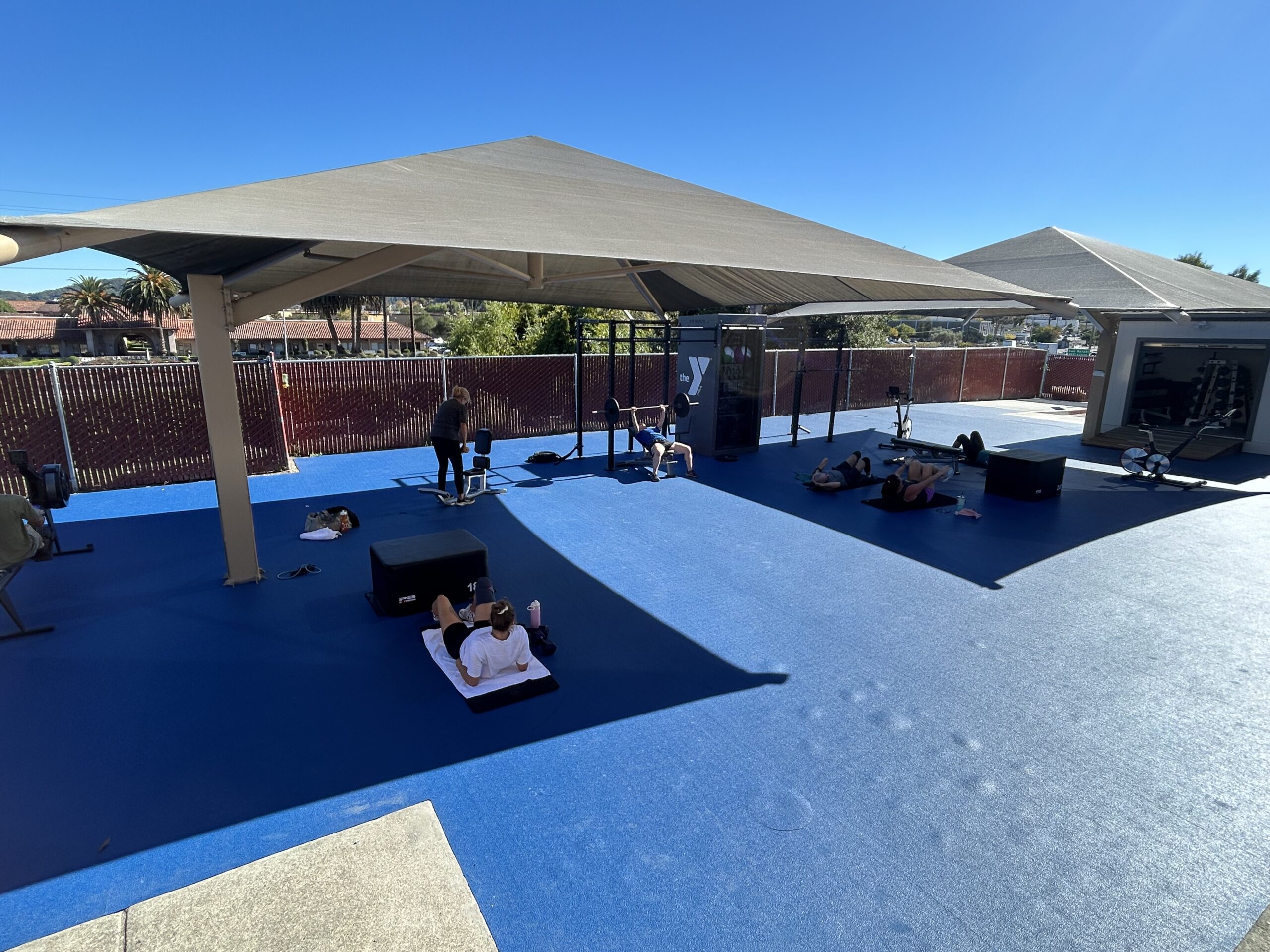 People are exercising on mats and equipment under a large outdoor canopy on a blue gym floor at the Marin YMCA, with clear skies and a fence framing this vibrant workout space.
