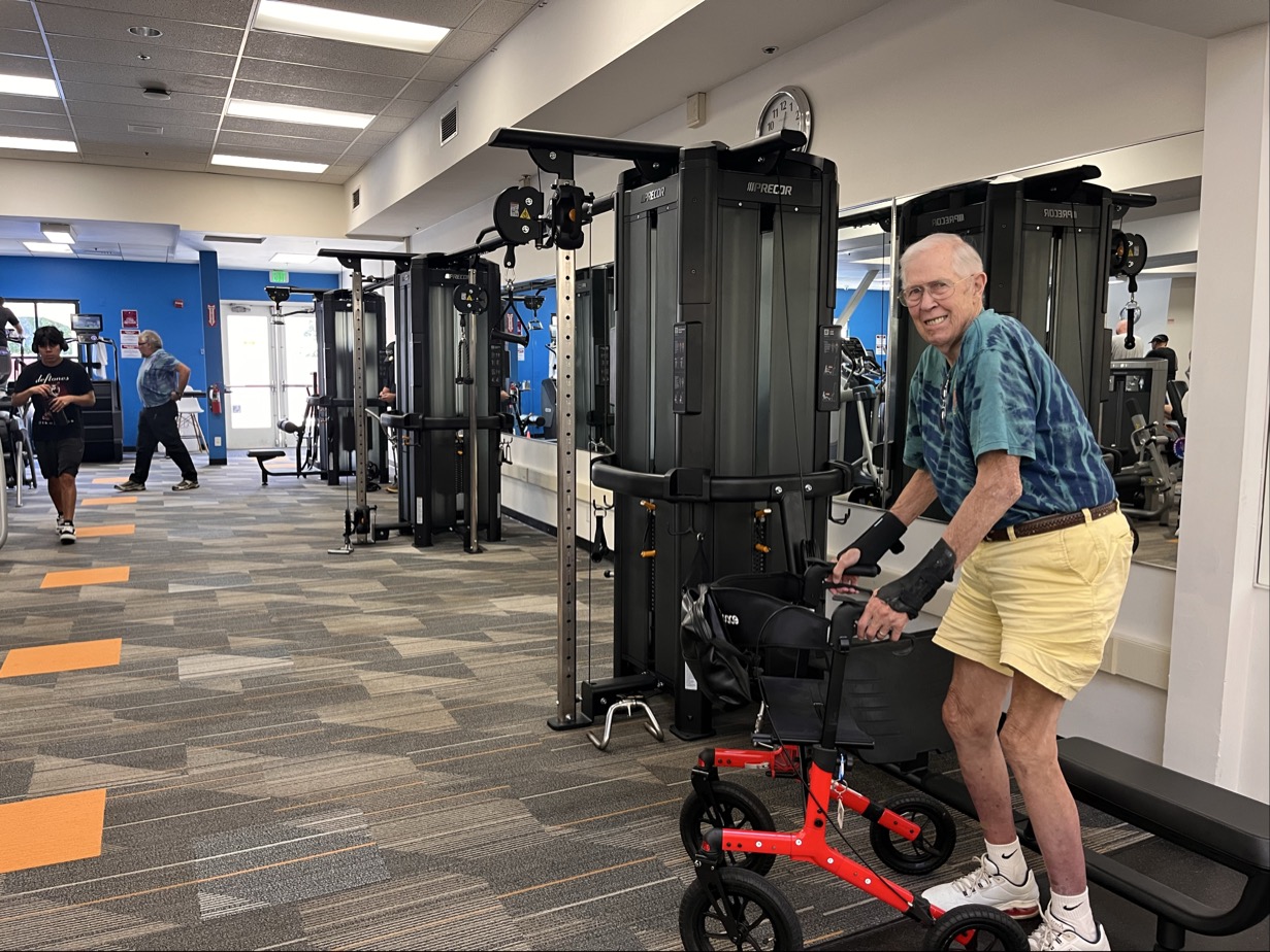 An older man with a striped shirt and yellow shorts uses a red walker in the Marin YMCA gym, surrounded by updated facilities, exercise machines, and others focused on their wellness.