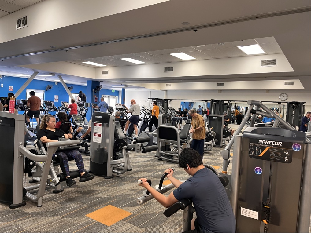 People are exercising on various gym machines in a busy YMCA fitness center with updated facilities, bright lighting, and modern equipment.