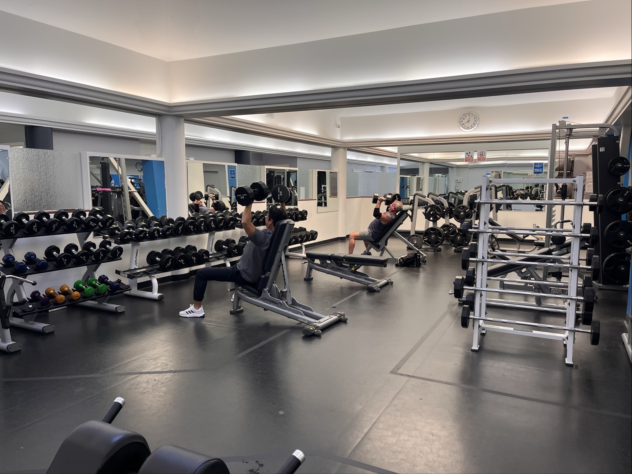 Two people are lifting dumbbells on benches in the updated facilities at Marin YMCA, surrounded by mirrors, free weights, and weight racks—a perfect setting to focus on wellness.