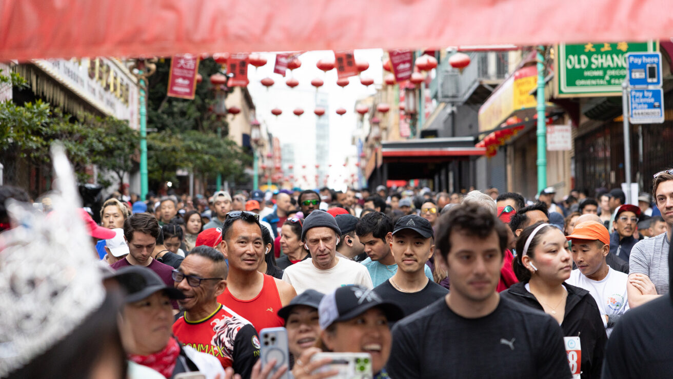 A large crowd of people walks through a city street decorated with red lanterns, some taking photos, during a public event or festival.