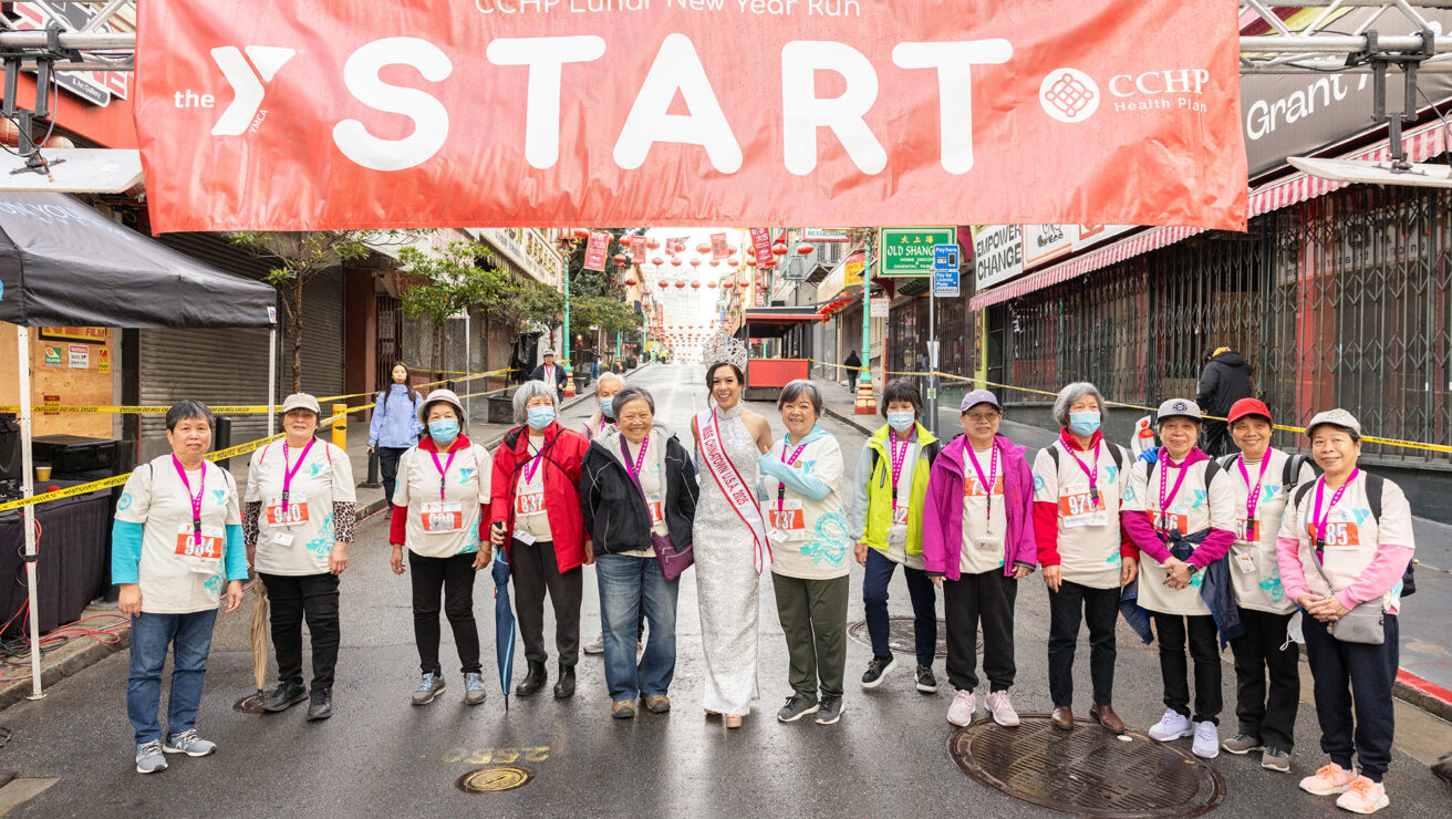 A group of people wearing race bibs and medals stand under a "START" banner for the CCHP Lunar New Year Run in an urban street setting.