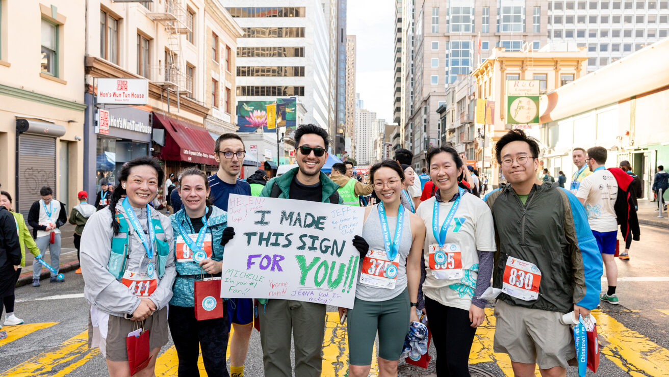 A group of marathon runners poses together on a city street; one person holds a sign that reads, “I MADE THIS SIGN FOR YOU!!” Tall buildings and other runners are visible in the background.