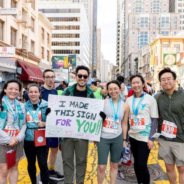 A group of marathon runners poses together on a city street; one person holds a sign that reads, “I MADE THIS SIGN FOR YOU!!” Tall buildings and other runners are visible in the background.