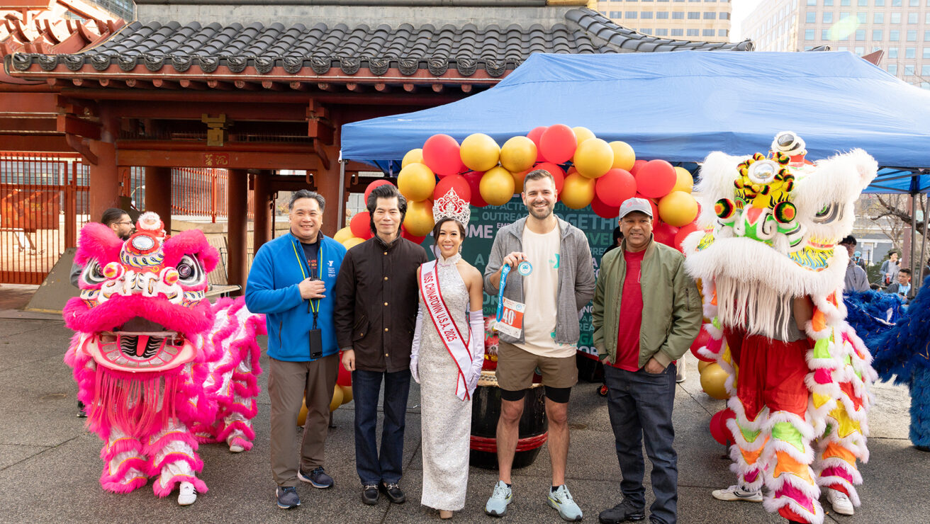 A group of people pose for a photo in front of a balloon arch, flanked by two lion dance costumes, at an outdoor cultural event in an urban setting.