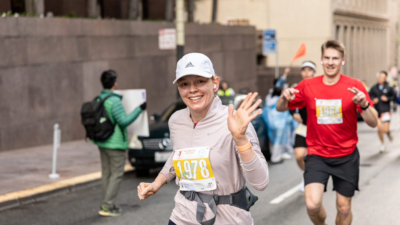 A woman wearing a white cap and race bib 1978 runs in a street marathon, smiling and waving at the camera. Other runners and spectators are visible in the background.