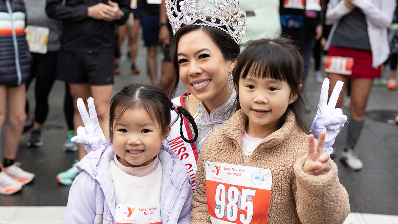 A woman wearing a crown and sash poses with two young girls showing peace signs, all wearing race bibs, at a street event with other participants in the background.