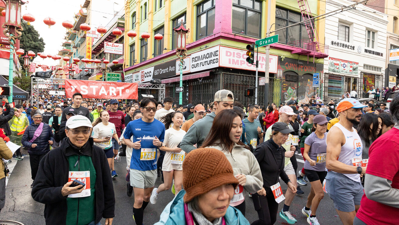 Runners participate in a race, passing under a red "START" banner on a city street lined with colorful buildings and lanterns. Spectators line the sidewalk.