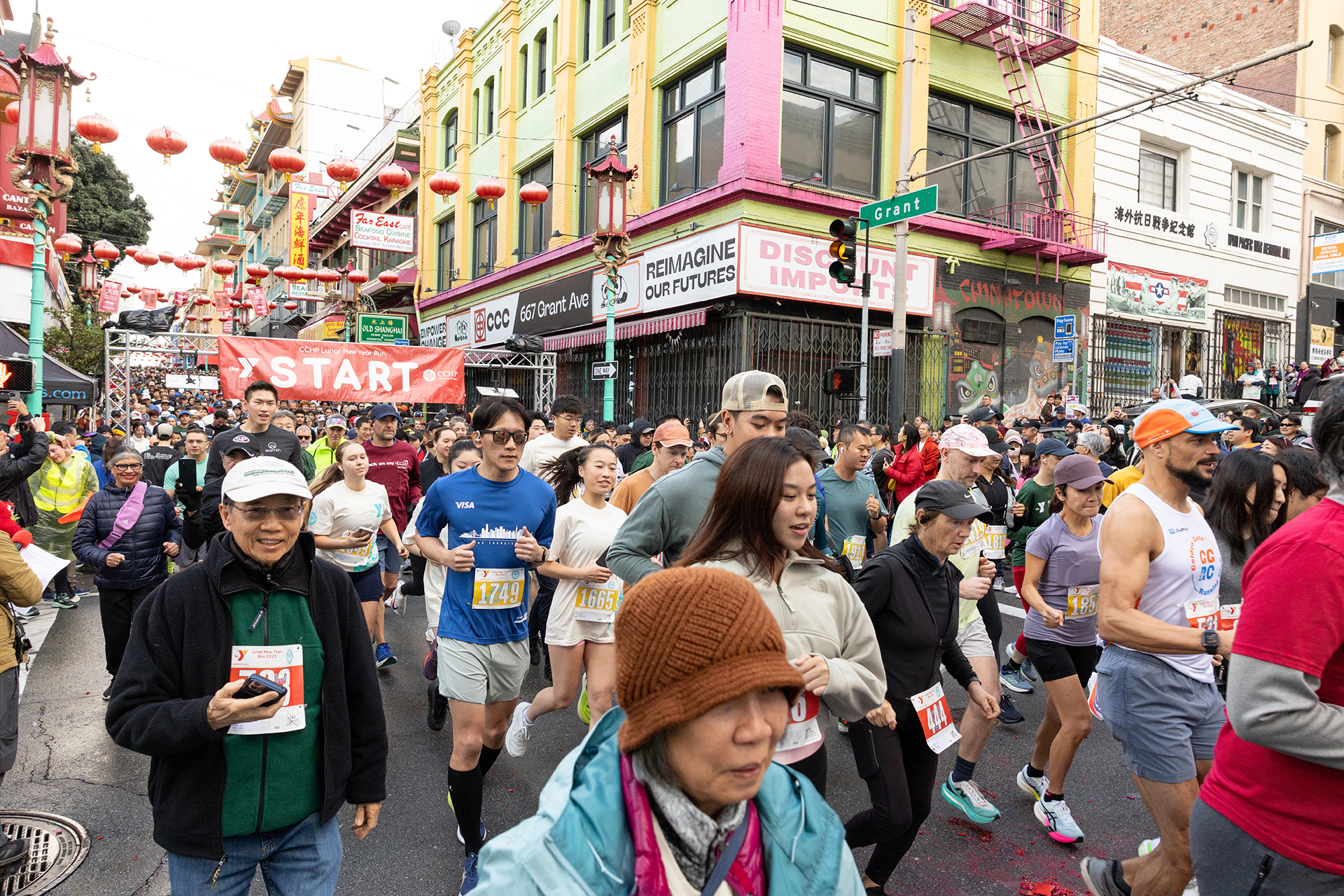 Runners participate in a race, passing under a red "START" banner on a city street lined with colorful buildings and lanterns. Spectators line the sidewalk.