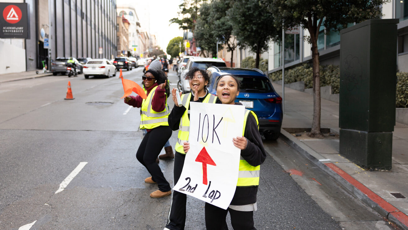 Three people in yellow safety vests stand on a city street, one holding a "10K 2nd Lap" sign, directing or cheering for an event.
