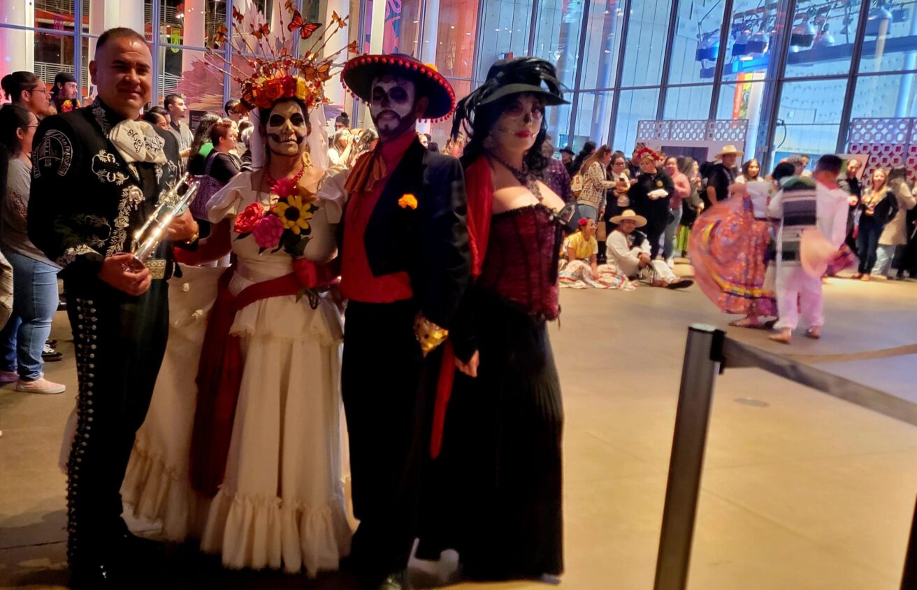 Four people in festive Day of the Dead costumes pose indoors at a crowded event, with colorful outfits and face paint.