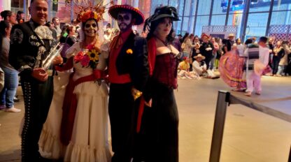 Four people in festive Day of the Dead costumes pose indoors at a crowded event, with colorful outfits and face paint.