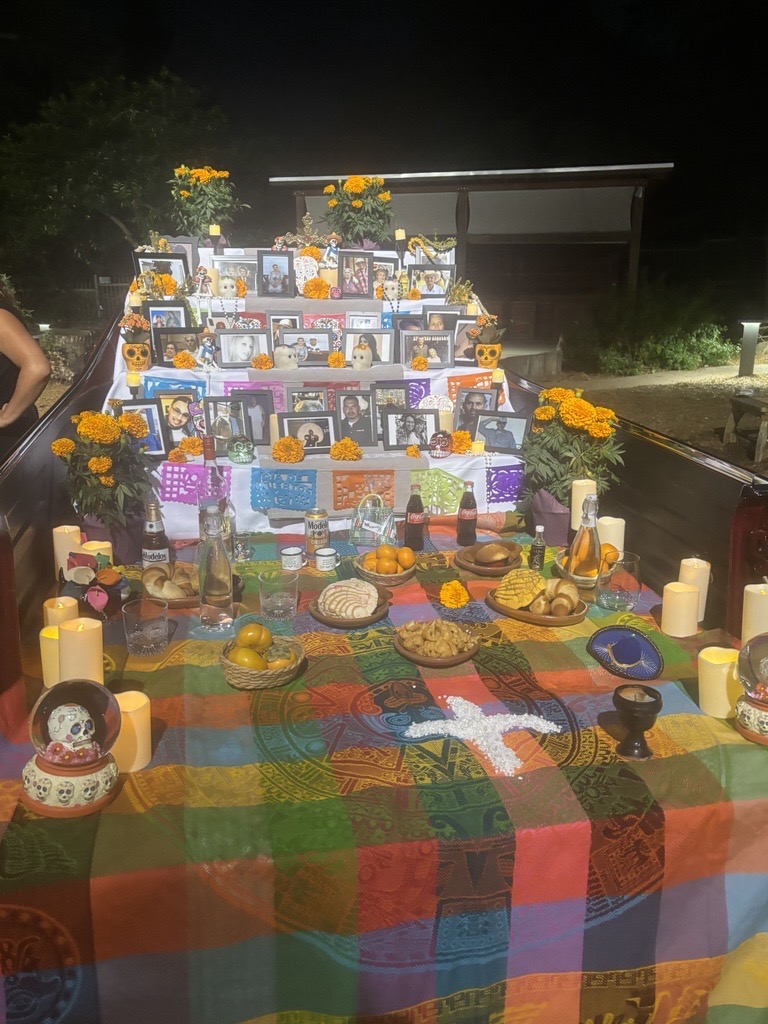A Día de los Muertos altar with framed photos, marigold flowers, candles, food, and drinks sits on a colorful tablecloth at night, honoring heritage and bringing community together.