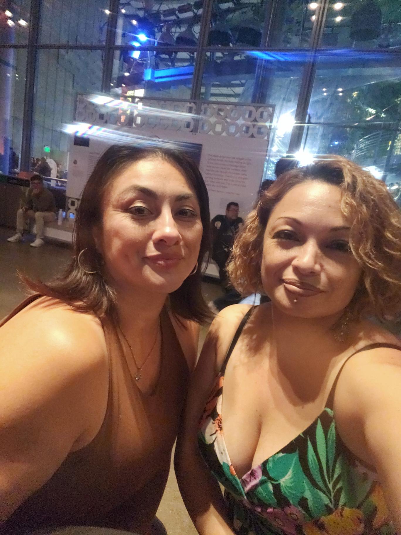 Two women pose for a selfie indoors with glass walls and several people seated in the background. Both are smiling slightly and dressed in sleeveless tops, celebrating their heritage in a warm community setting.
