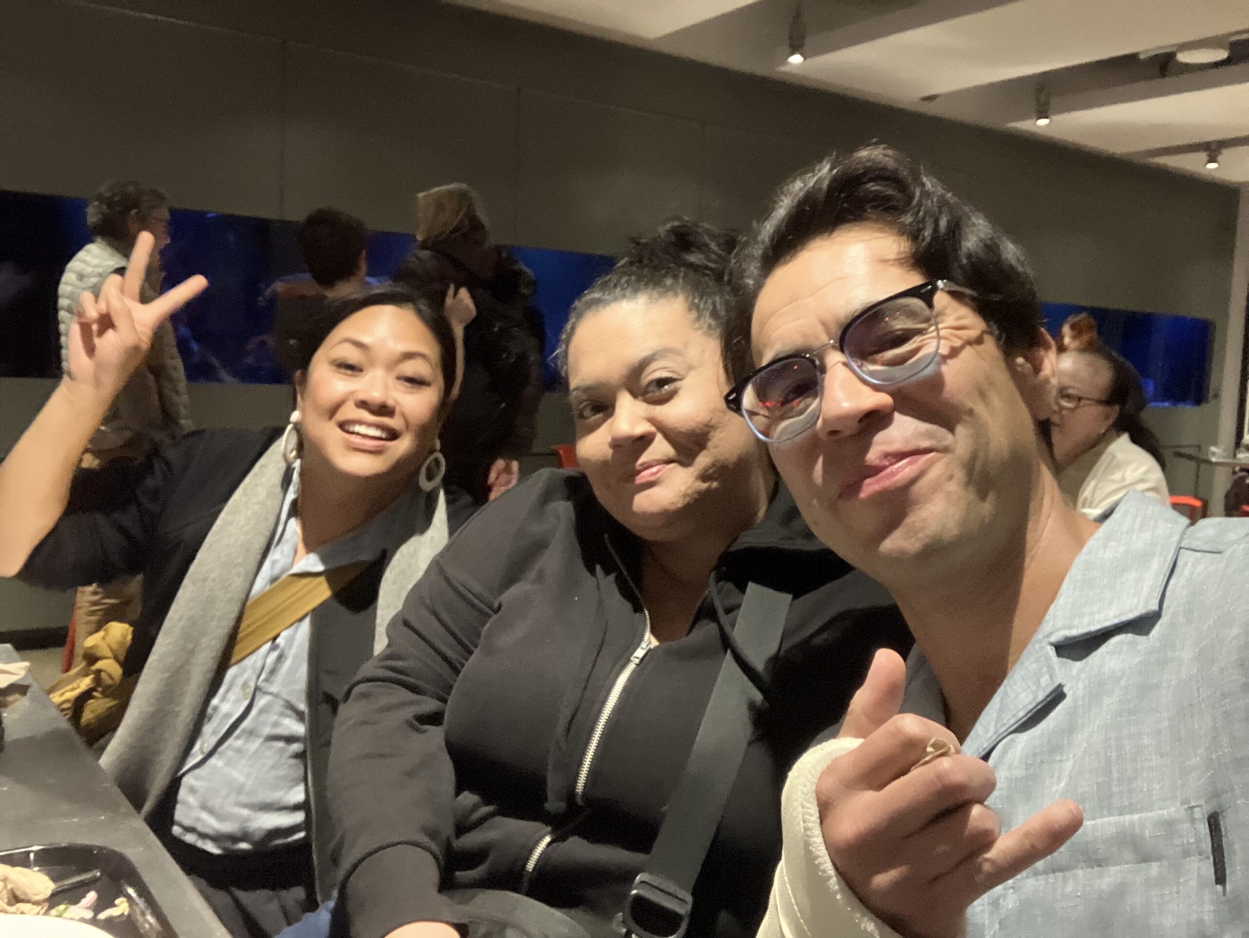 Three people sit together at a table indoors, smiling at the camera; one person is making a peace sign and another is making a shaka hand gesture, celebrating community and shared heritage.