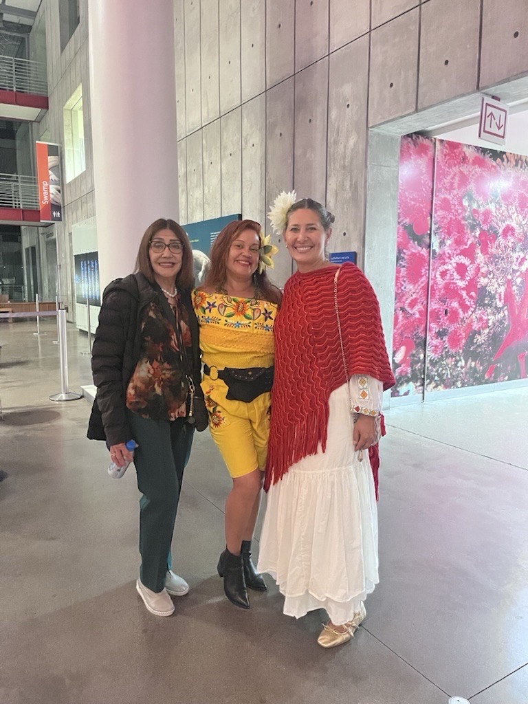 Three women pose indoors, celebrating community and heritage: one in a jacket and pants, another in a yellow folkloric outfit, and the third in a white dress with a red shawl and flower, evoking Día de los Muertos spirit.