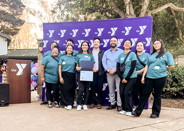 A group of people pose outdoors in front of a purple YMCA banner, with one person holding a certificate and a podium visible on the left.