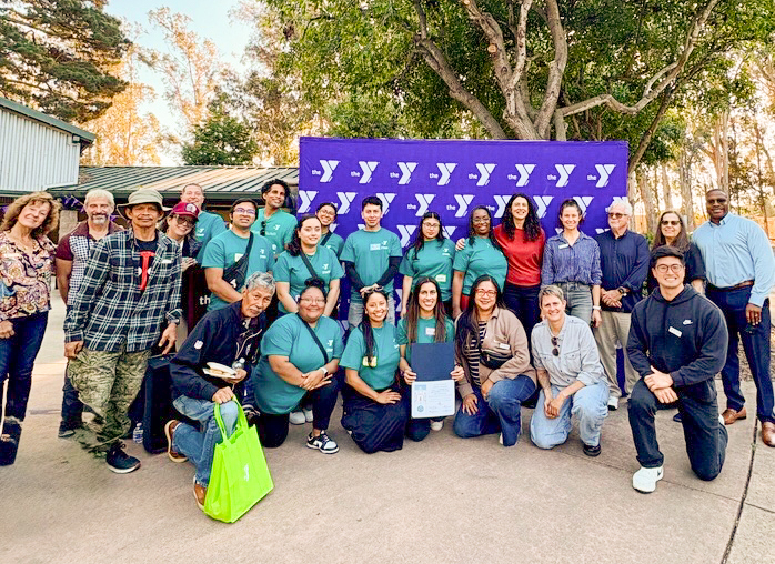 A group of people pose outdoors in front of a purple YMCA banner, with some standing and others kneeling, all smiling at the camera.