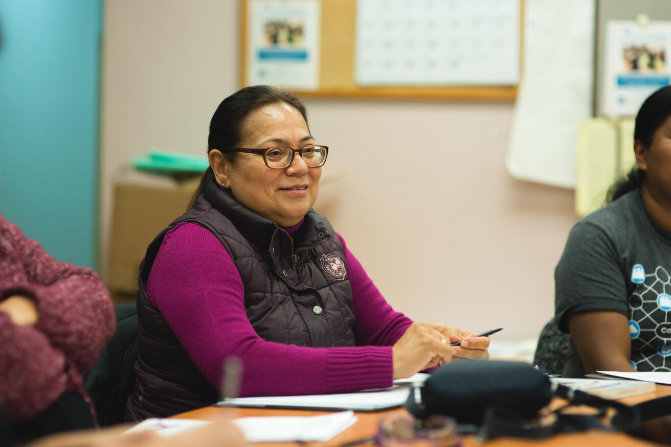 Woman with glasses and a maroon vest sits at a table holding a pen, with papers in front of her, in a classroom or meeting setting.