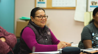 Woman with glasses and a maroon vest sits at a table holding a pen, with papers in front of her, in a classroom or meeting setting.