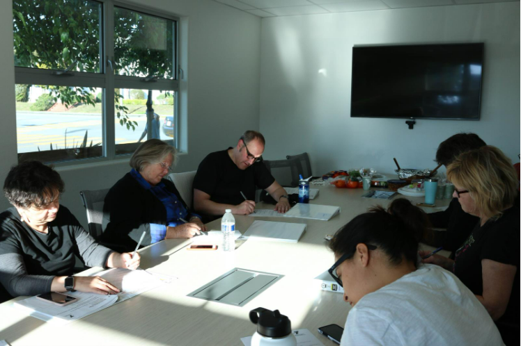Six people sit around a conference table, writing or reviewing documents related to diabetes prevention, with snacks and water bottles on the table in a well-lit meeting room for Diabetes Awareness Month.