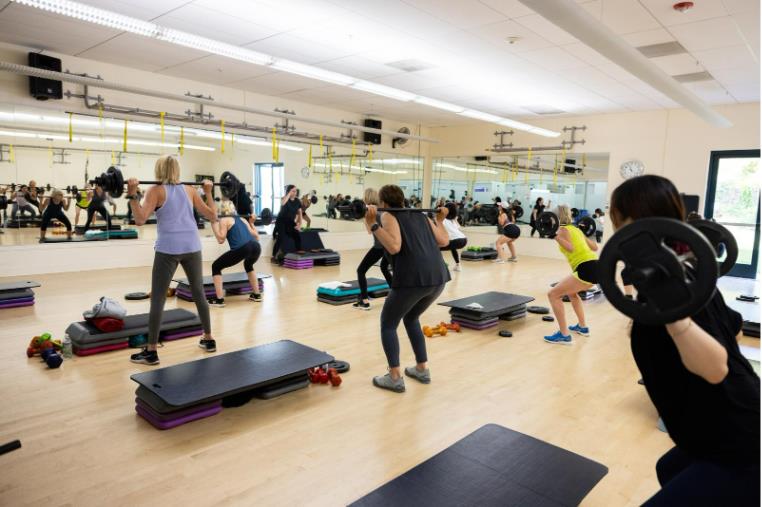 A group of people participate in a fitness class using barbells and aerobic steps in a mirrored exercise studio, promoting diabetes prevention during Diabetes Awareness Month.