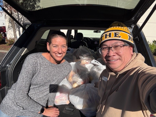 Two people sit in the back of a car filled with bags of groceries from the Marin YMCA’s December food distribution, smiling at the camera on a sunny day.