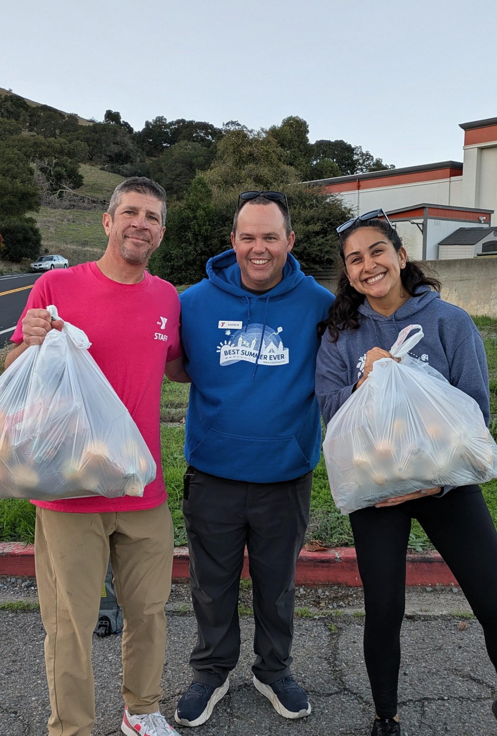 Three people stand outdoors in December, smiling as they hold large plastic bags filled with food items during a Marin YMCA food distribution. A hillside and buildings are visible in the background.
