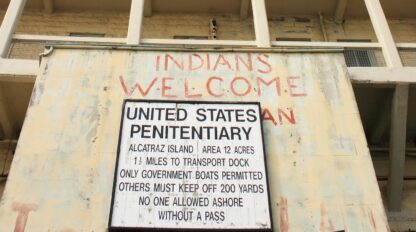 A building with a large sign reading "United States Penitentiary" and "Indians Welcome" painted in red above it, referencing Alcatraz Island and highlighting the enduring Indigenous Presence.