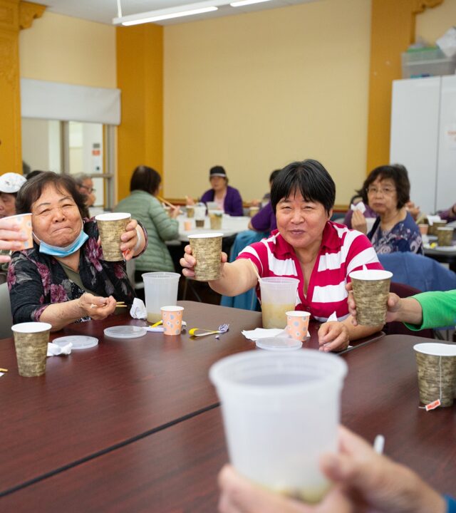 A group of seniors at the YMCA of Greater San Francisco sit around a table, raising cups in a toast during a communal meal in a brightly lit room.