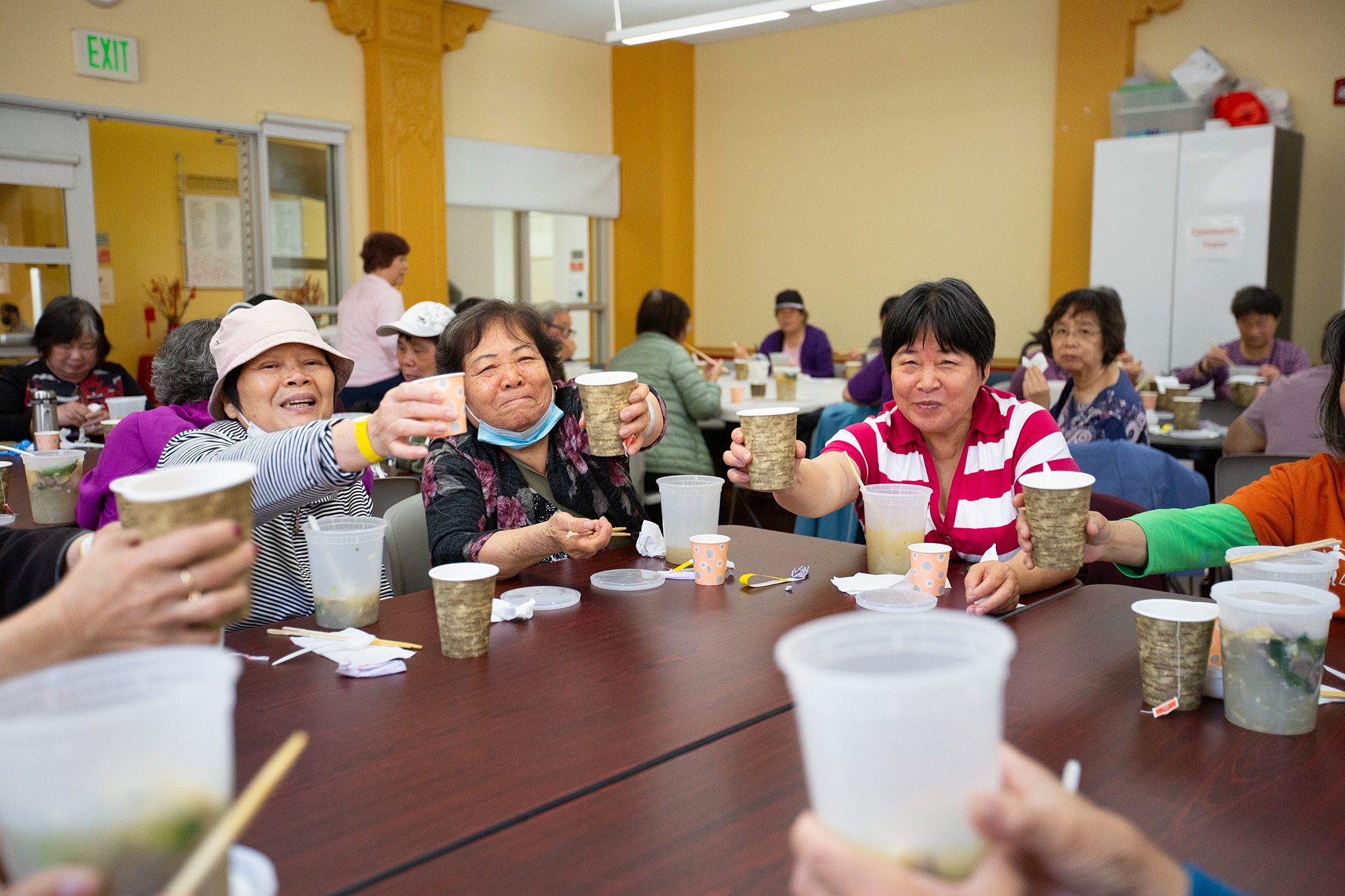 A group of seniors at the YMCA of Greater San Francisco sit around a table, raising cups in a toast during a communal meal in a brightly lit room.