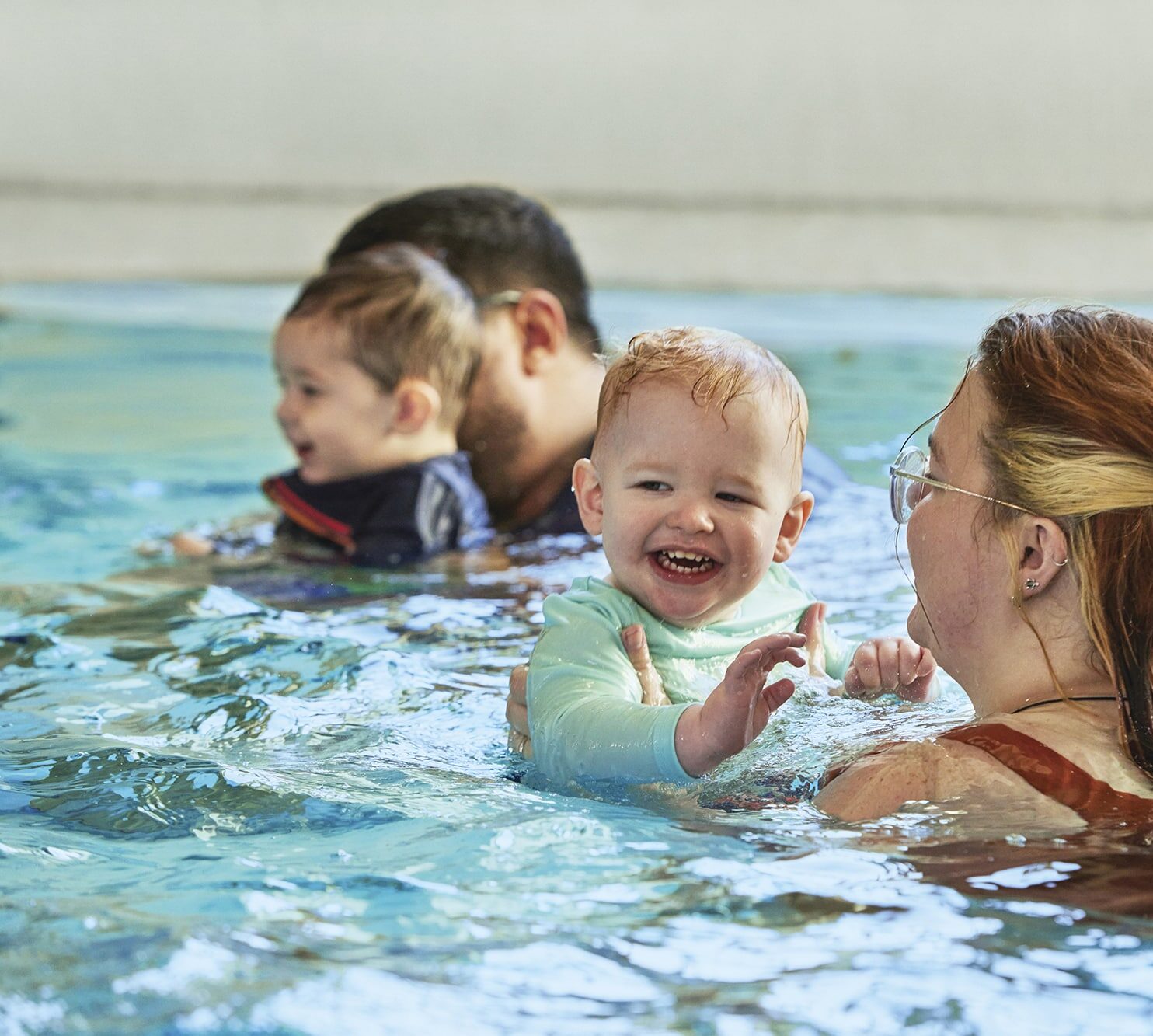 Two adults hold babies in a swimming pool. One baby smiles at the camera while being supported by a woman wearing glasses. The other adult and baby are in the background.