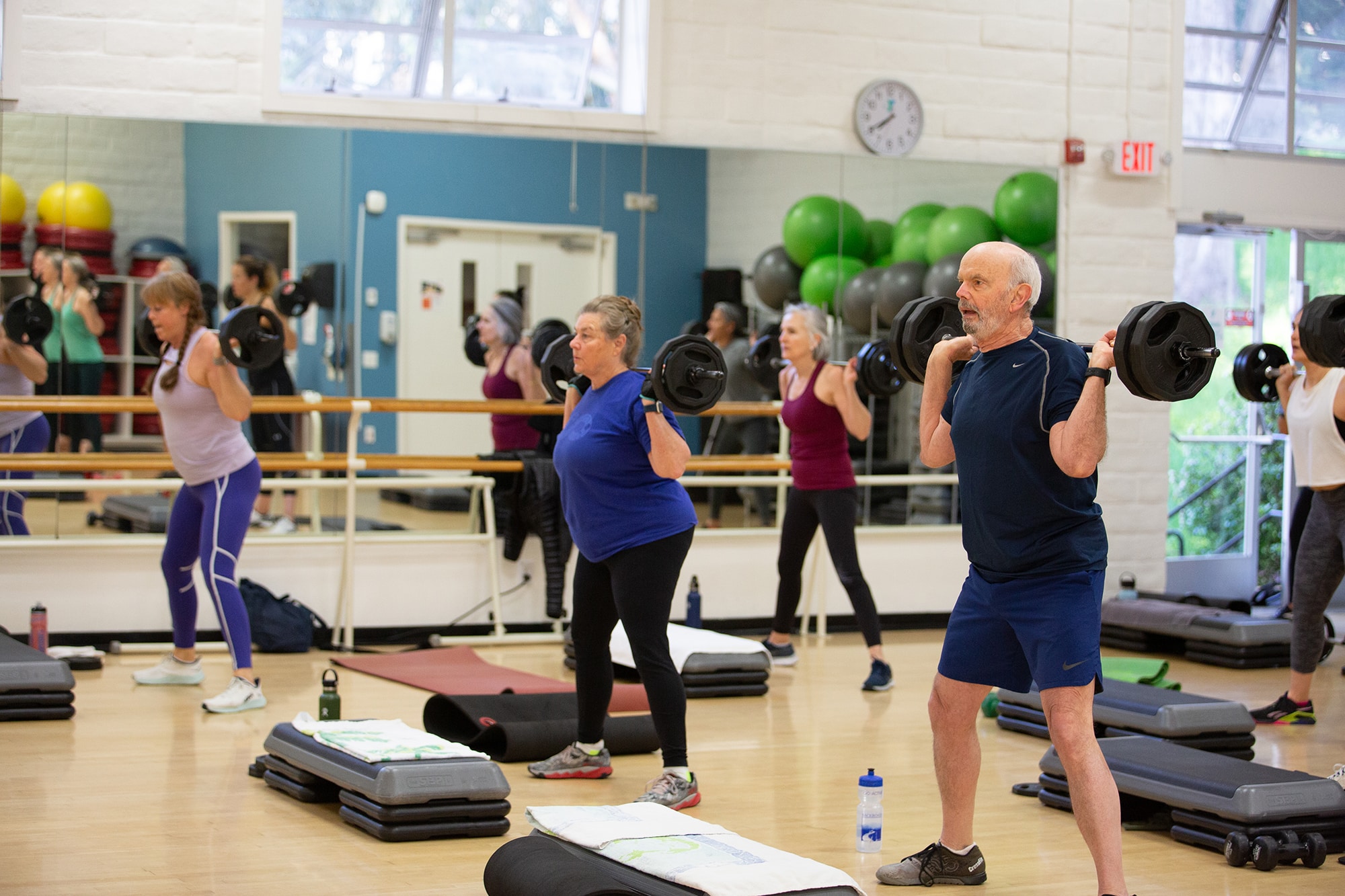 A group of older adults participate in a fitness class at the YMCA of Greater San Francisco, lifting barbells in a gym filled with mirrors, mats, and exercise equipment.
