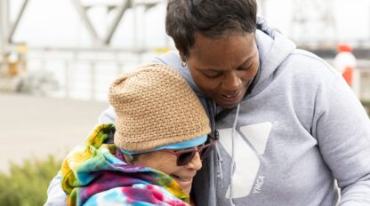 Two people wearing hoodies and winter accessories share a hug outdoors near a metal structure on a cloudy day, embodying the warmth and community spirit of the YMCA of Greater San Francisco.