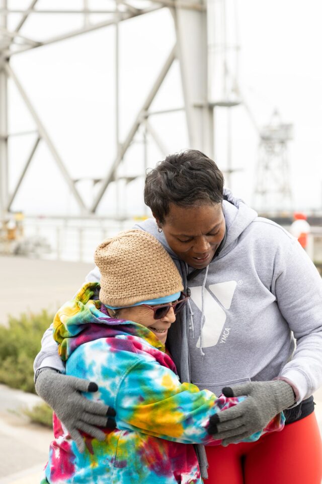 Two people wearing hoodies and winter accessories share a hug outdoors near a metal structure on a cloudy day, embodying the warmth and community spirit of the YMCA of Greater San Francisco.