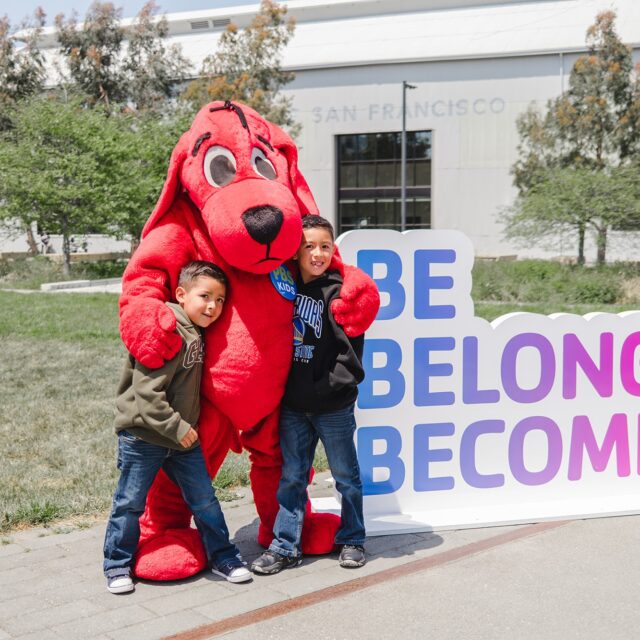 Two children pose with a person in a red dog costume next to a sign that reads "BE BELONG BECOME" at the YMCA of Greater San Francisco on a sunny day.