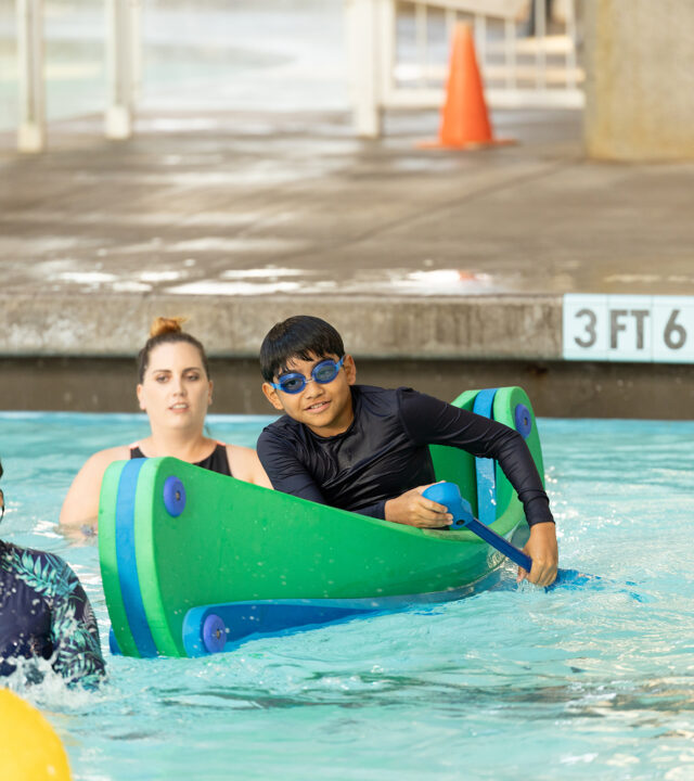 Three children in swimwear play in a swimming pool near a "3 FT 6 IN" depth marker, with pool toys and a supervising adult in the background during an annual report event.