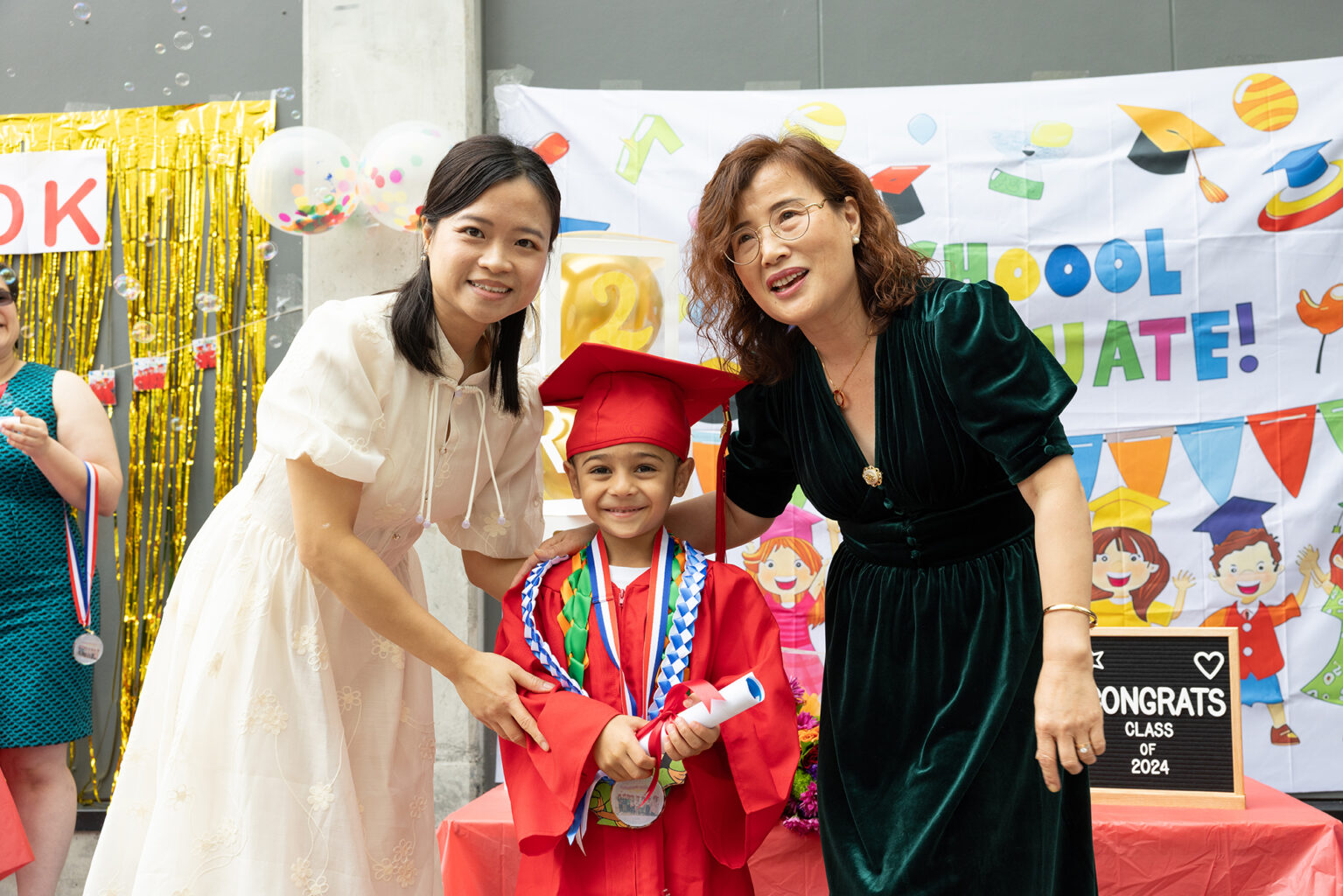 A young child in a red graduation gown and cap stands between two adults, smiling and holding a diploma, at the school’s Annual Report celebration.