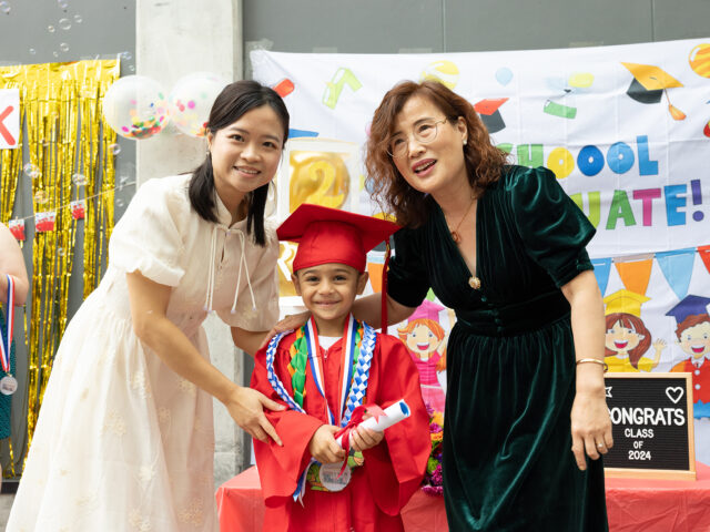 A young child in a red graduation gown and cap stands between two adults, smiling and holding a diploma, at the school’s Annual Report celebration.
