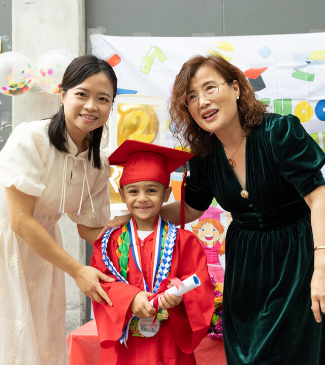 A young child in a red graduation gown and cap stands between two adults, smiling and holding a diploma, at the school’s Annual Report celebration.
