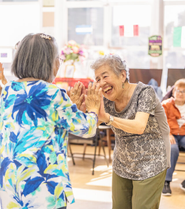 Two elderly women smile and clap hands together while others watch in a bright, communal room decorated with small flags—capturing the joyful spirit featured in our Annual Report.