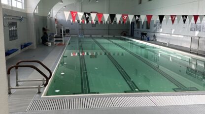 An indoor swimming pool at the Stonestown Family YMCA features clear water, lane dividers, and red, black, and white triangular flags above. Tools rest on the gray tile floor in the foreground.