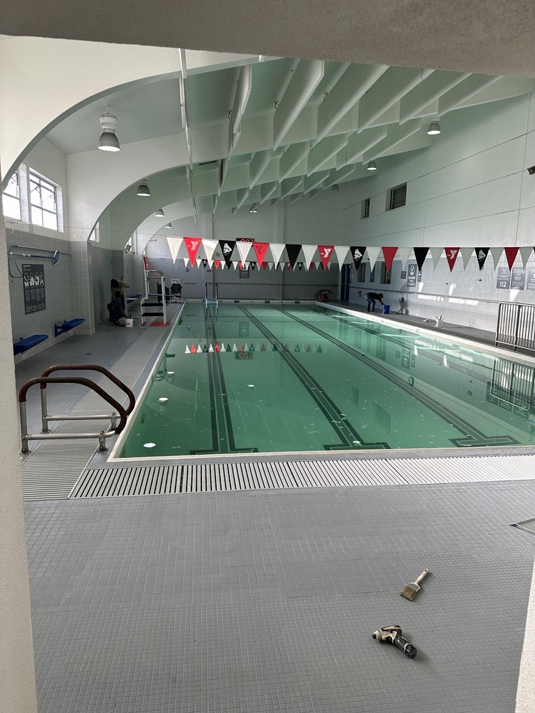 An indoor swimming pool at the Stonestown Family YMCA features clear water, lane dividers, and red, black, and white triangular flags above. Tools rest on the gray tile floor in the foreground.
