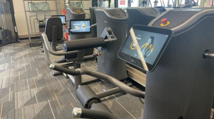 Row of modern strength training machines with digital screens displaying exercise instructions, set up in a well-lit Peninsula Family YMCA fitness center with carpeted flooring—ideal for Smart Strength Training routines.