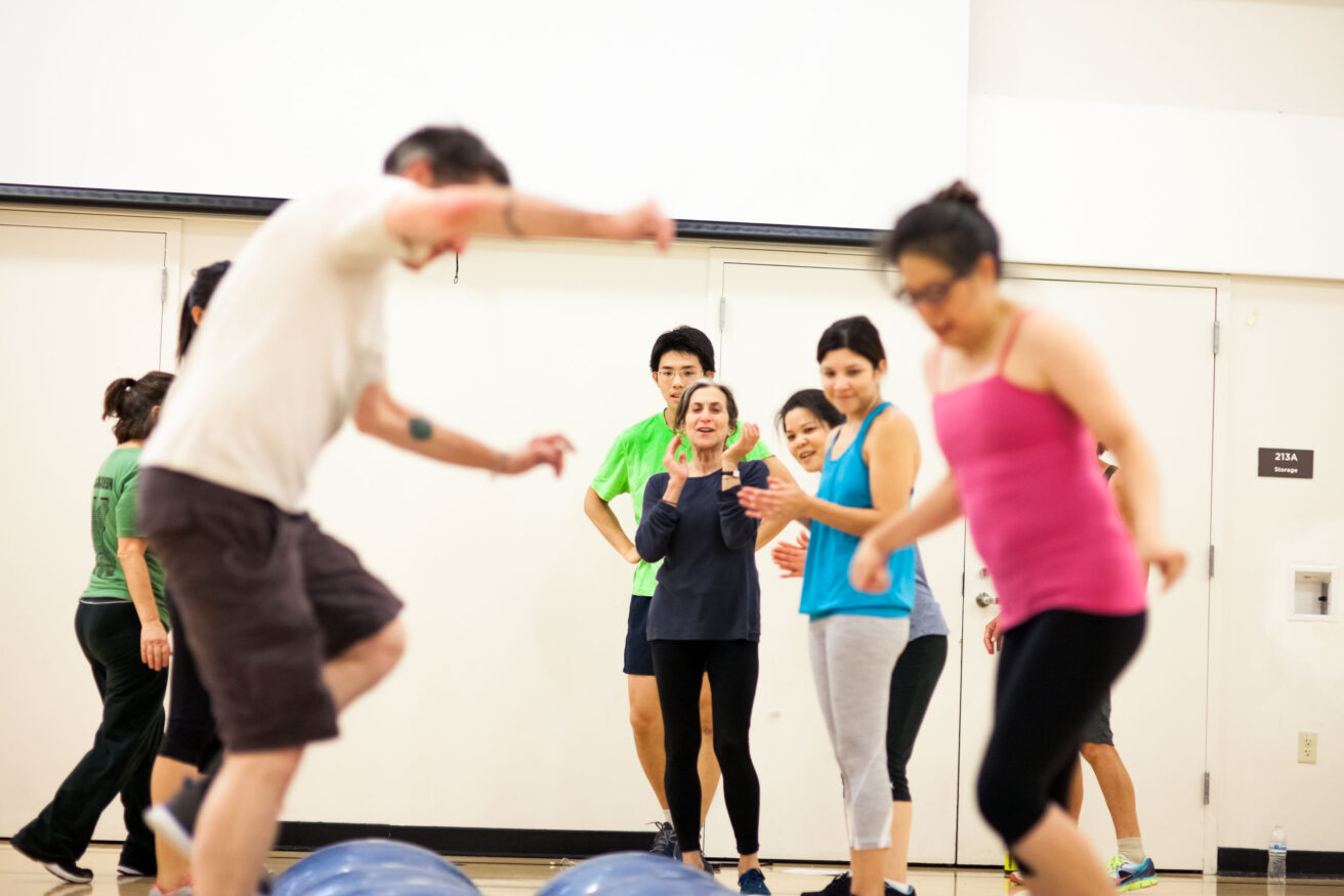 A group of adults participate in a fitness class indoors, with some balancing on exercise balls and others observing or waiting their turn.
