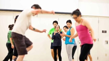 A group of adults participate in a fitness class indoors, with some balancing on exercise balls and others observing or waiting their turn.