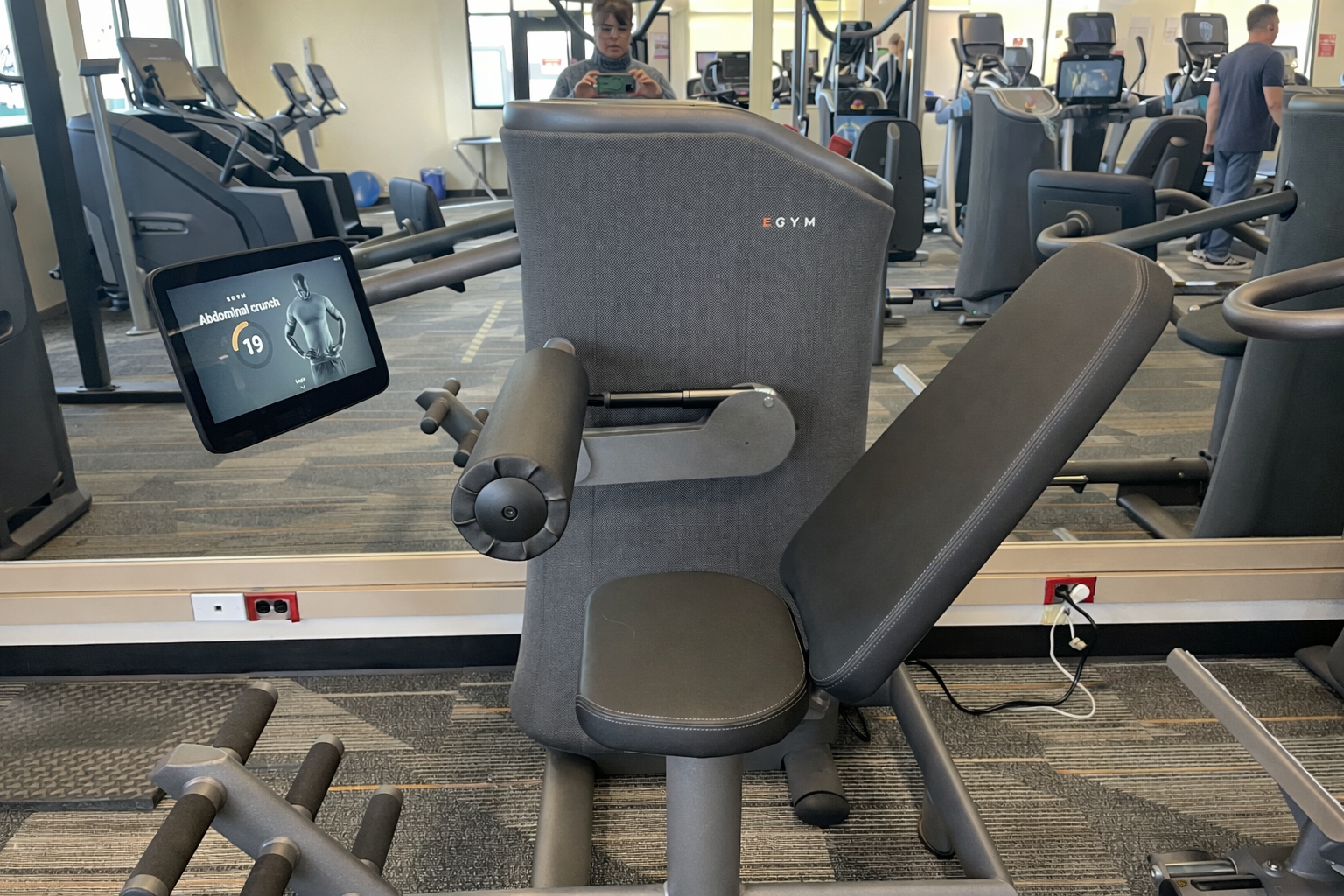 A seated leg extension machine with a digital display, part of the EGYM at Peninsula Family YMCA upgrade, is surrounded by other exercise equipment in the gym.